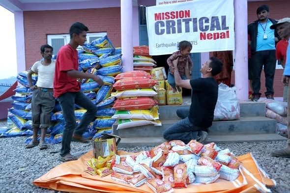 Volunteers stacking sacks of rice and packets of food outside a building, beneath a banner reading Mission Critical Nepal, Earthquake Relief 2015.